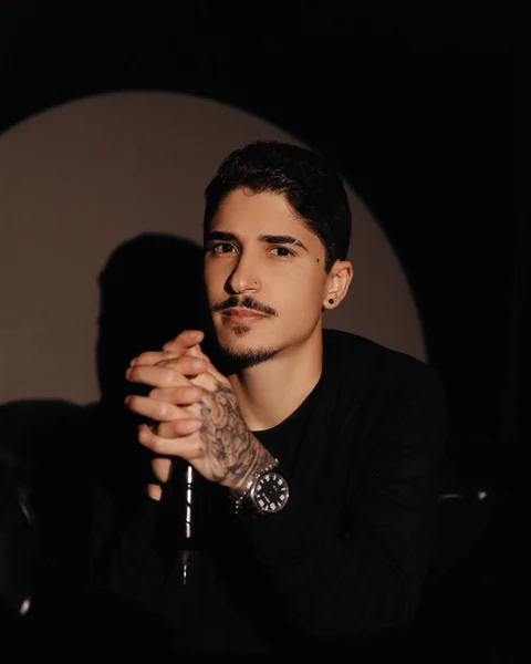 Jonatan Sanches sentado diante de um círculo de luz, de mãos entrelaçadas e com o antebraço tatuado à mostra.